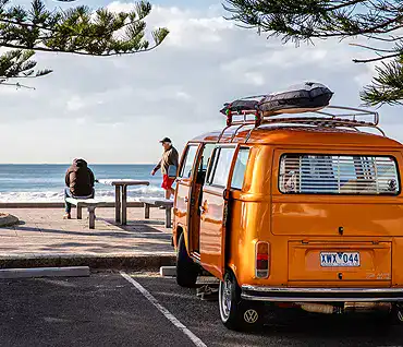 Orange vintage van parked near the beach with surfboard on top.